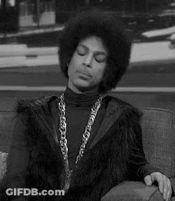 Prince Is Sitting On A Couch Wearing A Fur Vest And A Chain Around His Neck . GIF