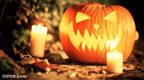 Pumpkin With Carved Face Next To Two Candles GIF
