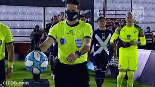 Referee With Face Mask Holding Soccer Ball In Front Of Players GIF