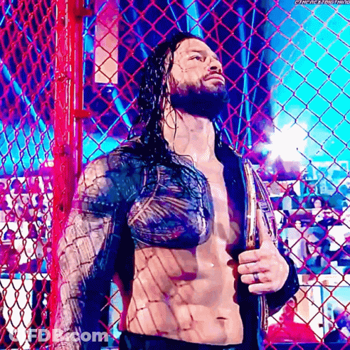 Roman Reigns Is Standing Behind A Chain Link Fence Holding A Belt . GIF