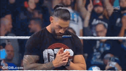 Roman Reigns Is Standing In A Wrestling Ring With His Hands Folded In Front Of A Crowd . GIF
