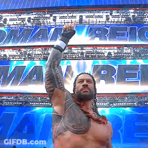 Roman Reigns Raises His Fist In The Air In Front Of A Banner That Says ' Roman Reigns ' GIF