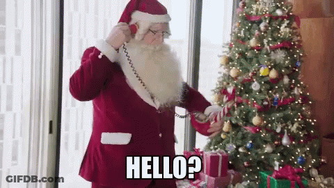 Santa Claus Is Talking On A Phone In Front Of A Christmas Tree And Says Hello ? GIF