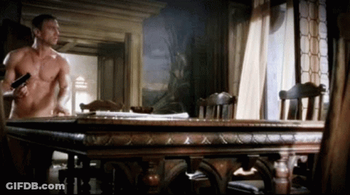 Shirtless Man Holding Gun In Front Of Wooden Table GIF