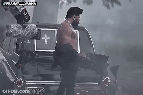 Shirtless Man Standing In Front Of Black Coffin With Cross GIF