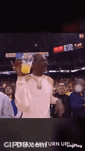 Snoop Dogg Is Holding A Cup Of Orange Juice In Front Of A Crowd Of People At A Basketball Game . GIF
