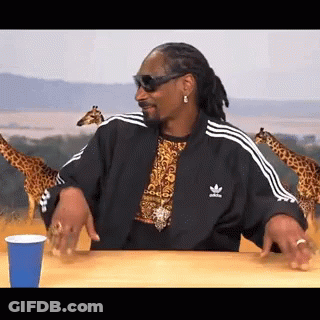 Snoop Dogg Is Sitting At A Table With A Blue Cup And Giraffes In The Background . GIF
