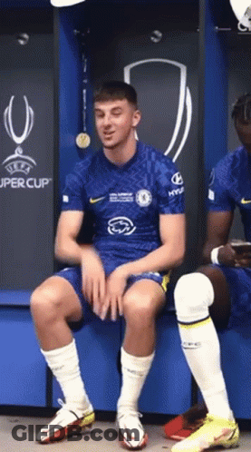 Soccer Player Sitting On Bench In Locker Room In Front Of UEFA Super Cup Sign GIF
