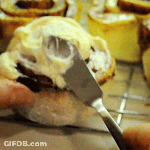 Someone Is Spreading Frosting On A Cinnamon Roll With A Knife GIF