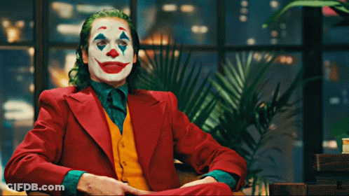 The Joker Is Sitting In A Chair With His Hands Folded GIF