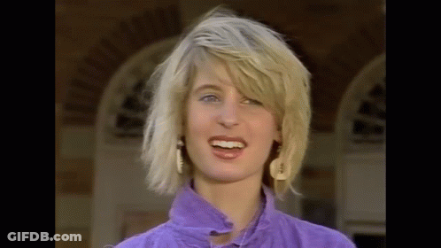 The Woman Is Wearing A Purple Shirt And Earrings And Is Smiling . GIF