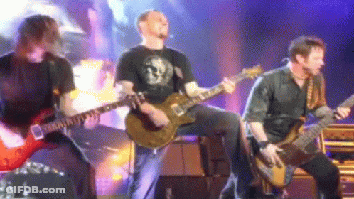 Three Men Are Playing Guitars On A Stage And One Of Them Is Wearing A Black Shirt With A Skull On It GIF