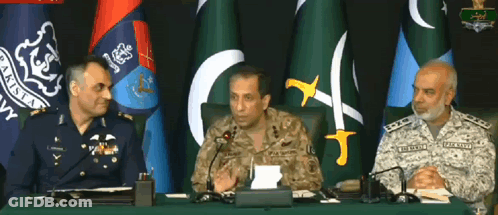 Three Men In Military Uniforms Sit At A Table With Microphones In Front Of Flags GIF