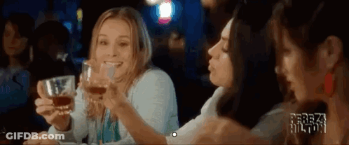 Three Women Are Sitting At A Table Holding Glasses Of Whiskey And Smiling . GIF
