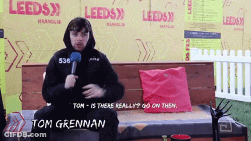 Tom Grennan Sitting On Bench With Microphone In Front Of Leeds Sign GIF