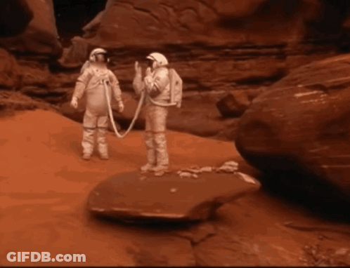 Two Astronauts Standing Next To Each Other On A Rock In The Desert GIF