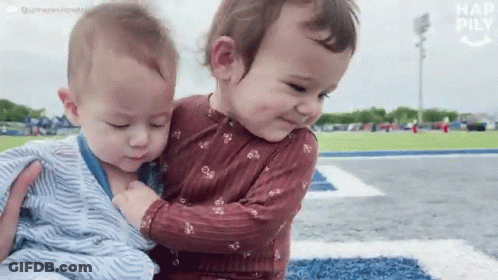 Two Babies Are Hugging Each Other On A Field And One Of Them Is Wearing A Striped Shirt GIF
