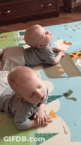 Two Babies Laying On A Mat With A Giraffe On It And A Box That Says Ozo On It GIF