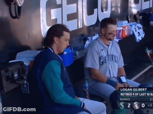 Two Baseball Players Sitting In A Dugout With Logan Gilbert On The Screen GIF