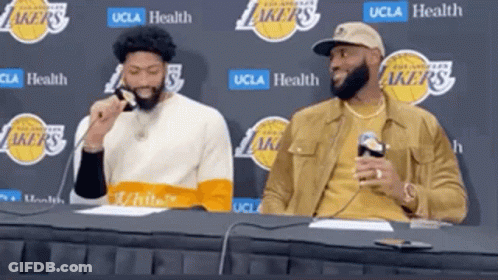 Two Basketball Players Are Sitting At A Table With A Lakers Logo Behind Them GIF