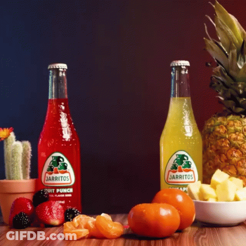 Two Bottles Of Jarritos Sit On A Wooden Table Surrounded By Fruit GIF