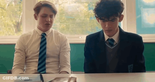 Two Boys Are Sitting At A Desk In A Classroom . GIF