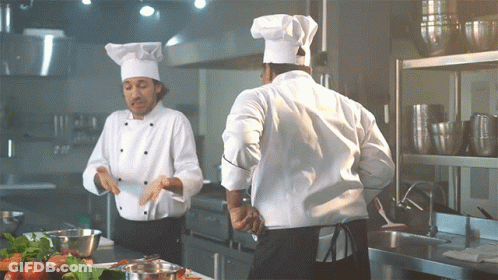 Two Chefs In Kitchen, One In White Hat GIF