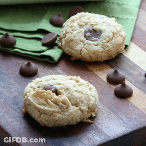 Two Cookies On A Wooden Cutting Board With Chocolate Chips Around Them GIF
