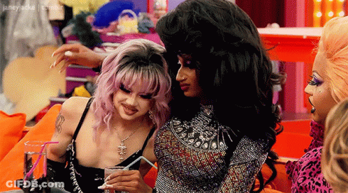 Two Drag Queens Sitting On Couch One Holding Straw GIF