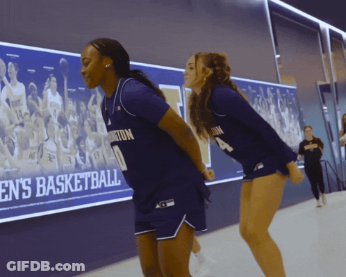 Two Female Basketball Players In Front Of Men's Basketball Wall GIF