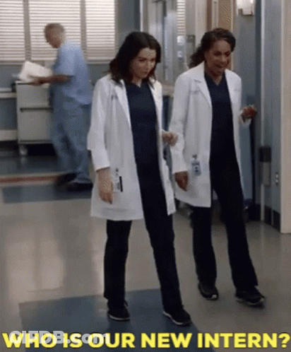 Two Female Doctors Are Walking Down A Hallway With The Words " Who Is Our New Intern " Behind Them GIF