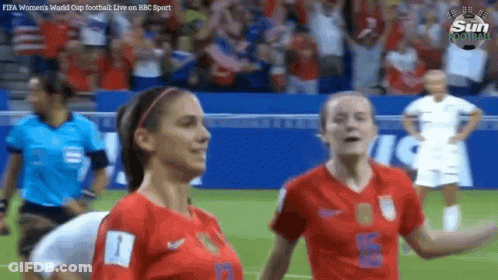 Two Female Soccer Players On A Field With A Referee In The Background GIF