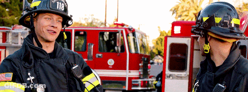Two Firefighters Are Talking In Front Of A Fire Truck With The Number 118 On The Helmet GIF