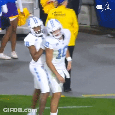 Two Football Players Are Hugging Each Other On The Field While A Cheerleader Stands Behind Them . GIF
