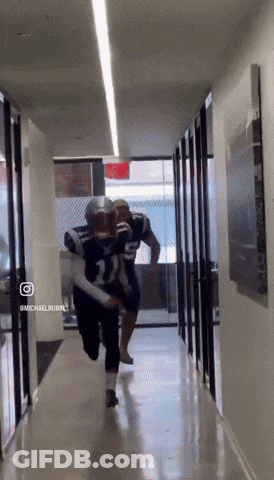 Two Football Players Are Running Down A Hallway With A Red Exit Sign Behind Them GIF