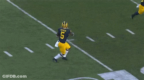 Two Football Players Are Running On The Field With The Letter M In The Background GIF