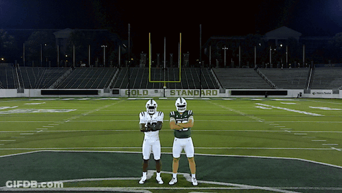 Two Football Players Standing On A Field With A Sign That Says Gold Standard In The Background GIF