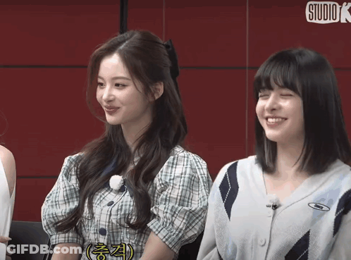 Two Girls Are Sitting Next To Each Other And Smiling In Front Of A Red Wall That Says Studio K GIF
