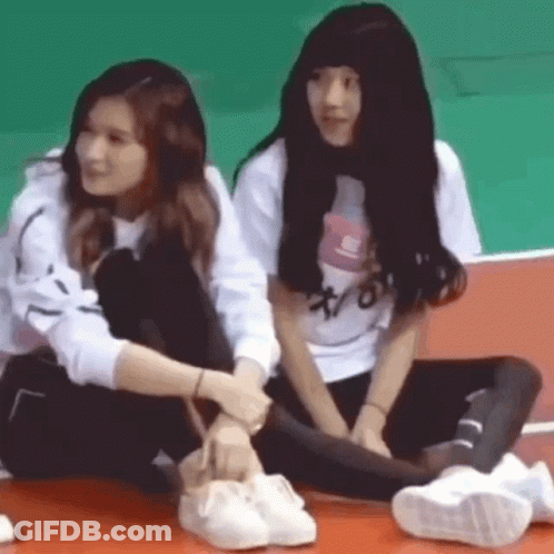 Two Girls Are Sitting Next To Each Other On The Floor And One Of Them Is Tying Another Girl 'S Shoe . GIF