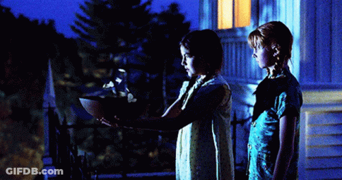 Two Girls Are Standing In Front Of A House At Night And One Is Holding A Bowl GIF