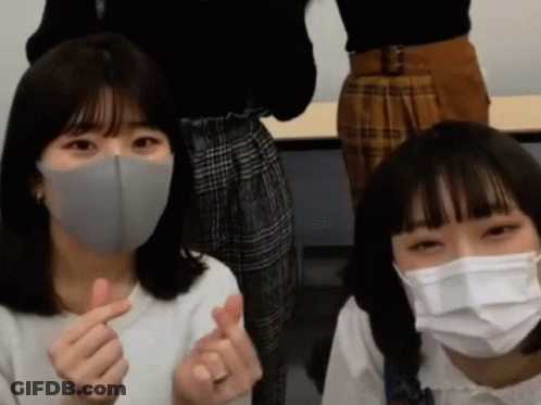 Two Girls Wearing Face Masks Are Making A Heart Shape With Their Fingers GIF