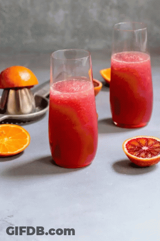 Two Glasses Of Red Juice Next To Oranges And A Juice Press GIF