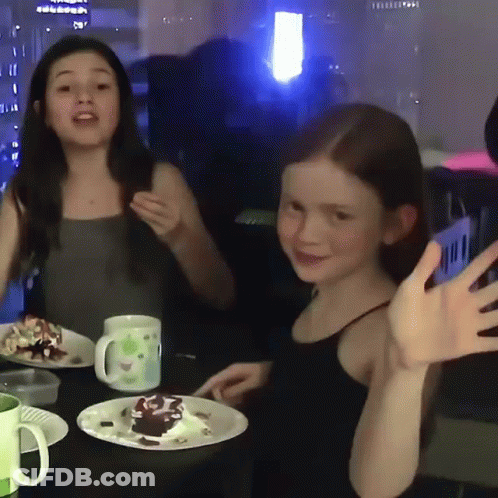 Two Little Girls Are Sitting At A Table With Plates Of Food GIF