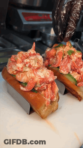 Two Lobster Rolls Are Being Prepared On A Cutting Board By A Person Wearing Black Gloves GIF