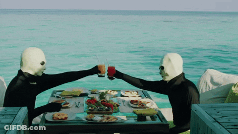 Two Masked People Toasting At A Table With Food GIF