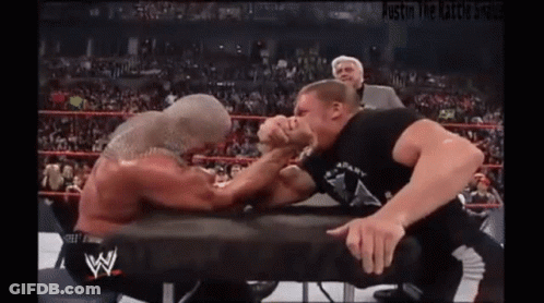 Two Men Are Arm Wrestling In A Wrestling Ring With A Referee Watching . GIF