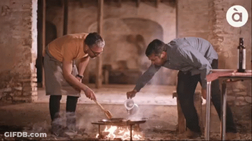 Two Men Are Cooking Over A Fire With The Letter A On The Bottom Right GIF