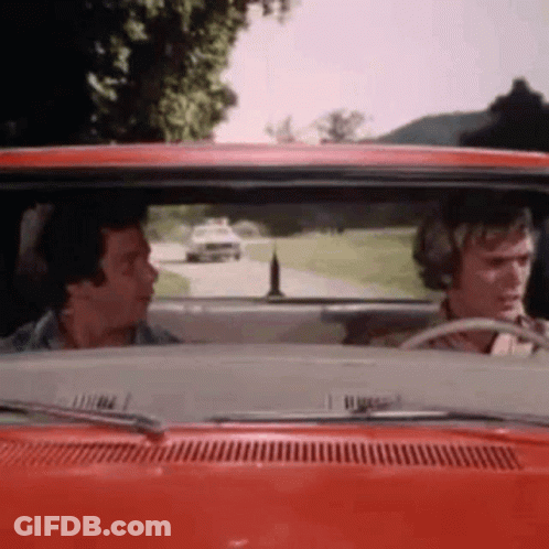 Two Men Are Driving A Red Car Down A Road . GIF