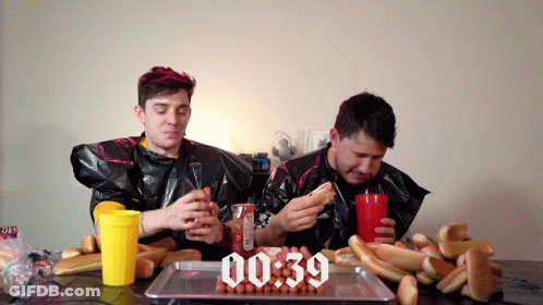 Two Men Are Eating Hot Dogs And Drinking Soda In Front Of A Tray Of Hot Dogs That Says 00:39 On It GIF