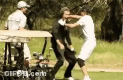 Two Men Are Fighting On A Golf Course While A Golf Cart Is Behind Them . GIF